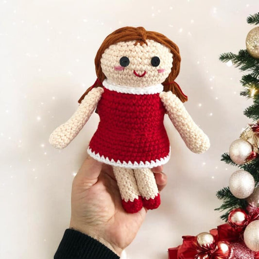 A crocheted version of the doll from misfit island being held up next to a Christmas tree. 