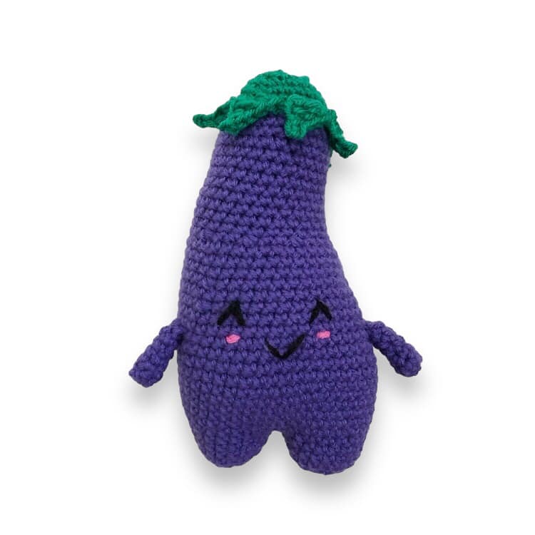 A crocheted doll resembling a cute eggplant with a smiling face standing against a white background. 