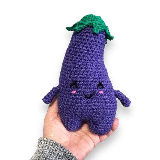 Hand holding a crochet eggplant with a purple body, green leaf, and smiling with pink cheeks. 