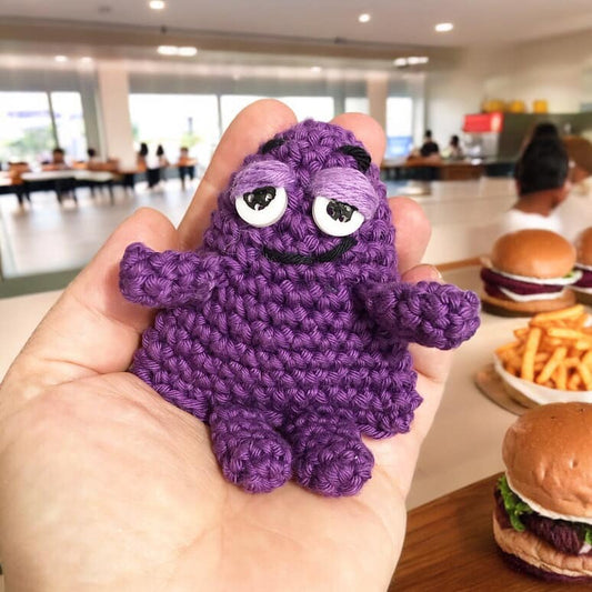 A small crochet toy resembling Grimace being held in a person's hand inside a burger joint. 