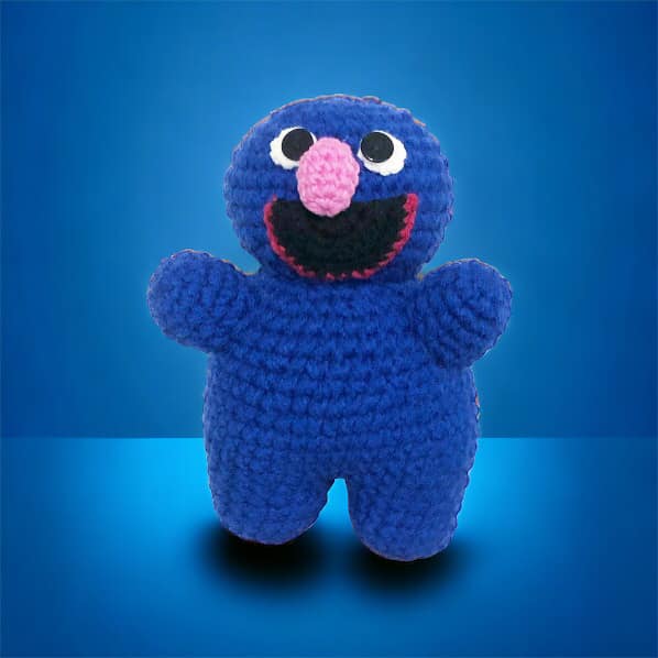 A crocheted version of Grover from Sesame Street standing against a blue background. 