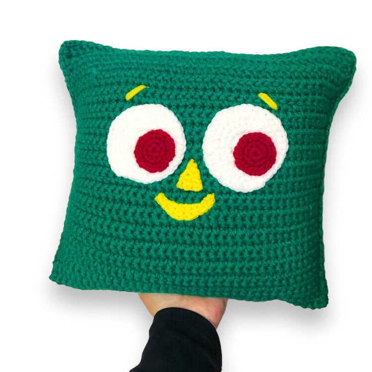 A crocheted pillow of Gumby's face being held up against a white background. 