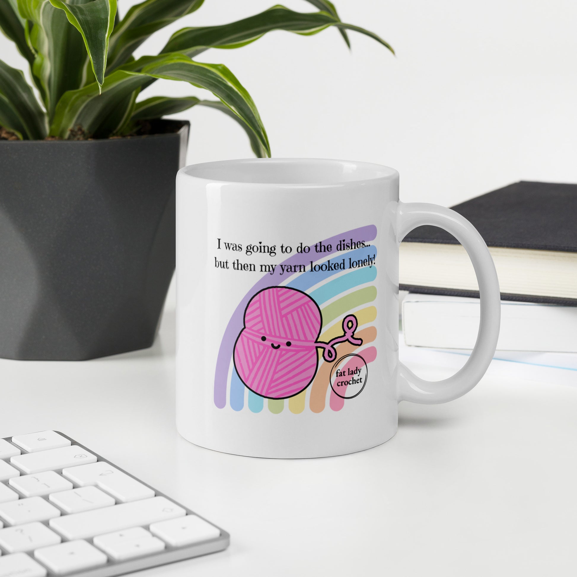 White mug with smiling pink yarn against pastel rainbow on a white desk with a an apple keyboard and potted plant