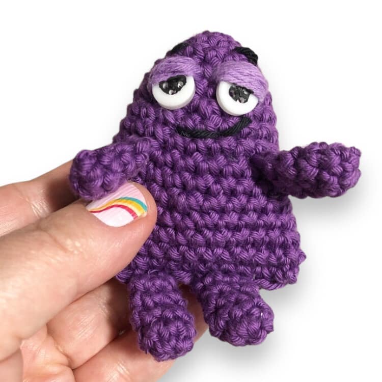 A tiny crochet doll resembling Grimace being held in a hand against a white background.
