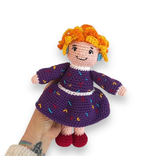 A crocheted doll of Mrs. Frizzle being held by a hand against a white background. 