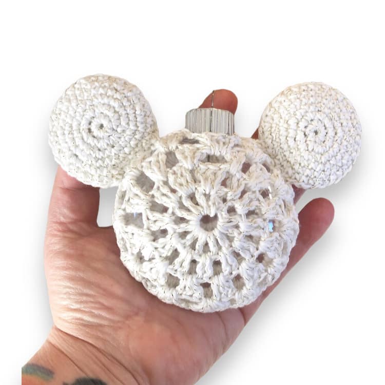 A crocheted white Mickey Mouse ornament being held up against a white background. 