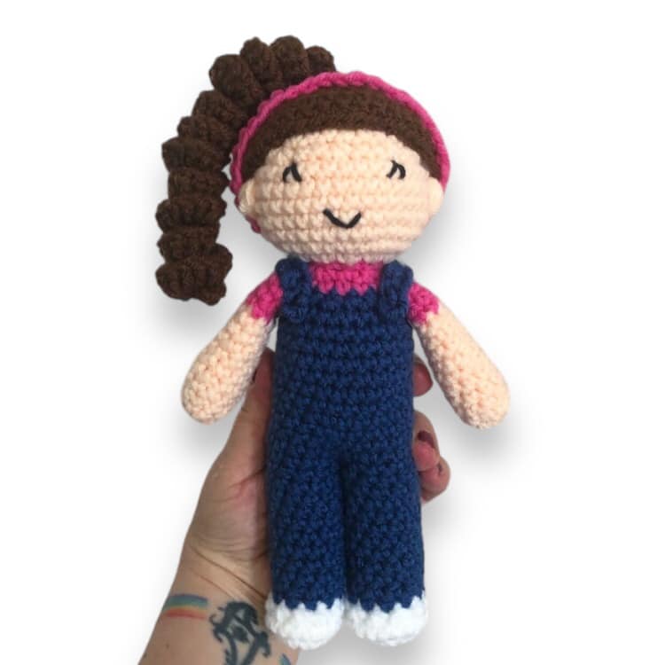 A crocheted doll resembling the kids content creator Ms. Rachel being held up against a white background. 
