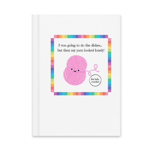 White journal with the quote "I was going to do the dishes...but my yard looked lonely!" on it with a pink smiling yarn ball set inside a rainbow border. 