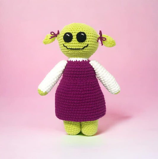 A crochet doll of Mona from Nanalan standing against a pink background. 