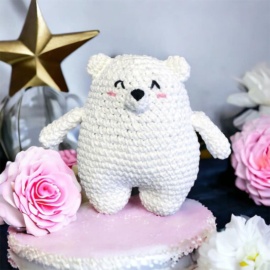 A crochet doll resembling a round polar bear standing amongst pink roses with a decorative gold star in the background. 