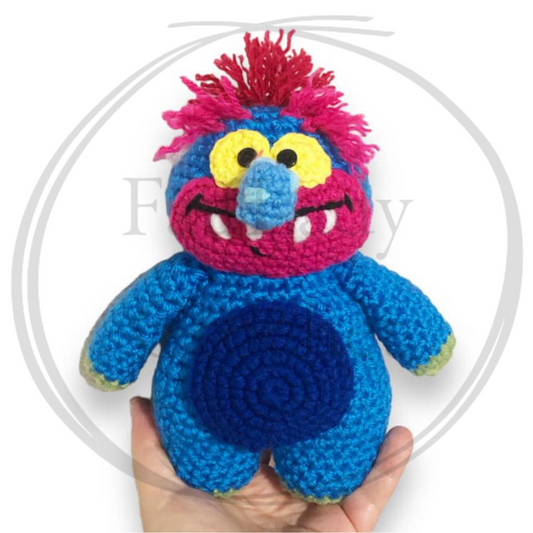A crocheted blue monster with a long wart nose, pink and blue face with fangs and pink hair being held up against a white background. 