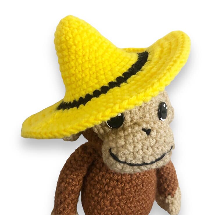 A crochet doll of Curious George wearing a crocheted yellow hat. 