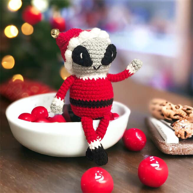 A crochet doll of a grey alien dressed as Santa Claus sitting on the edge of a bowl full of what I am assuming are maraschino cherries. 