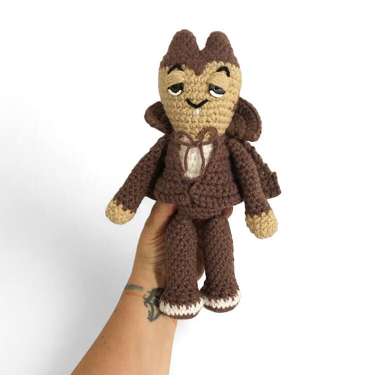 A crochet plushie resembling Count Chocula, with a brown body, white and beige accents, and small black buttons for eyes.