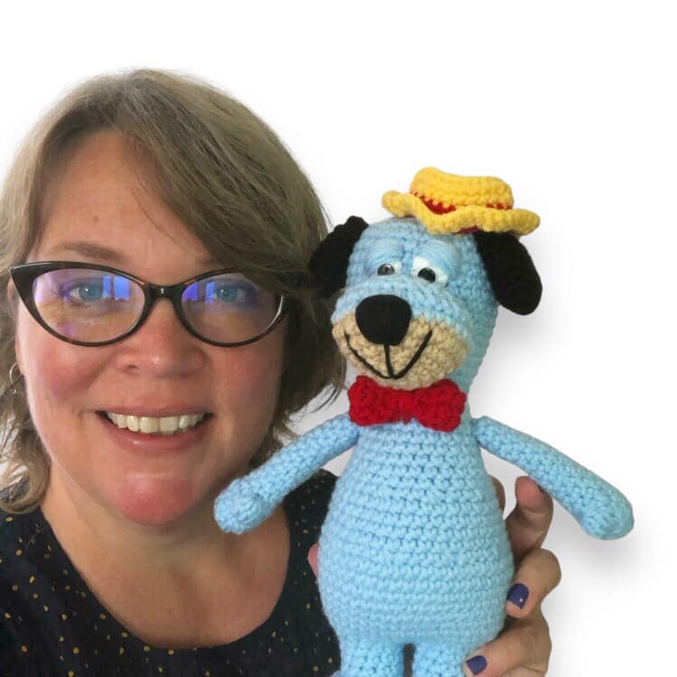 A woman smiling and holding up a crochet version of Huckleberry hound. 