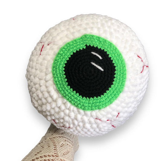 A large crocheted eyeball with a green iris being held up against a white background. 