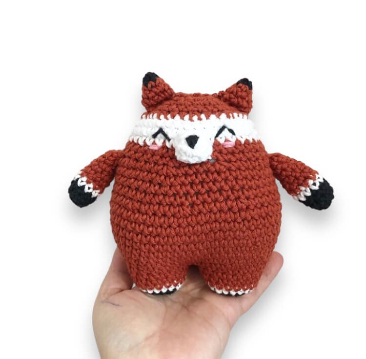 A crocheted doll resembling a chubby fox being held up against a white background. 
