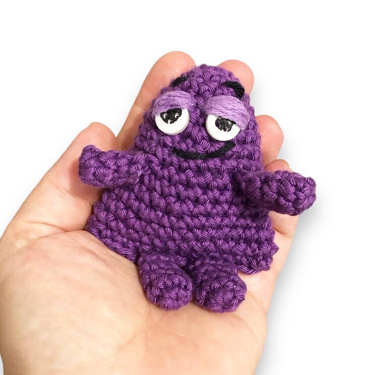 A tiny crochet doll resembling Grimace being held in a hand against a white background.