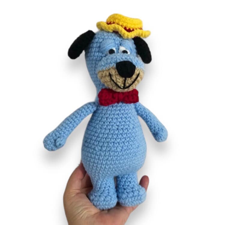 A crochet doll resembling Huckleberry Hound being held up against a white background. 