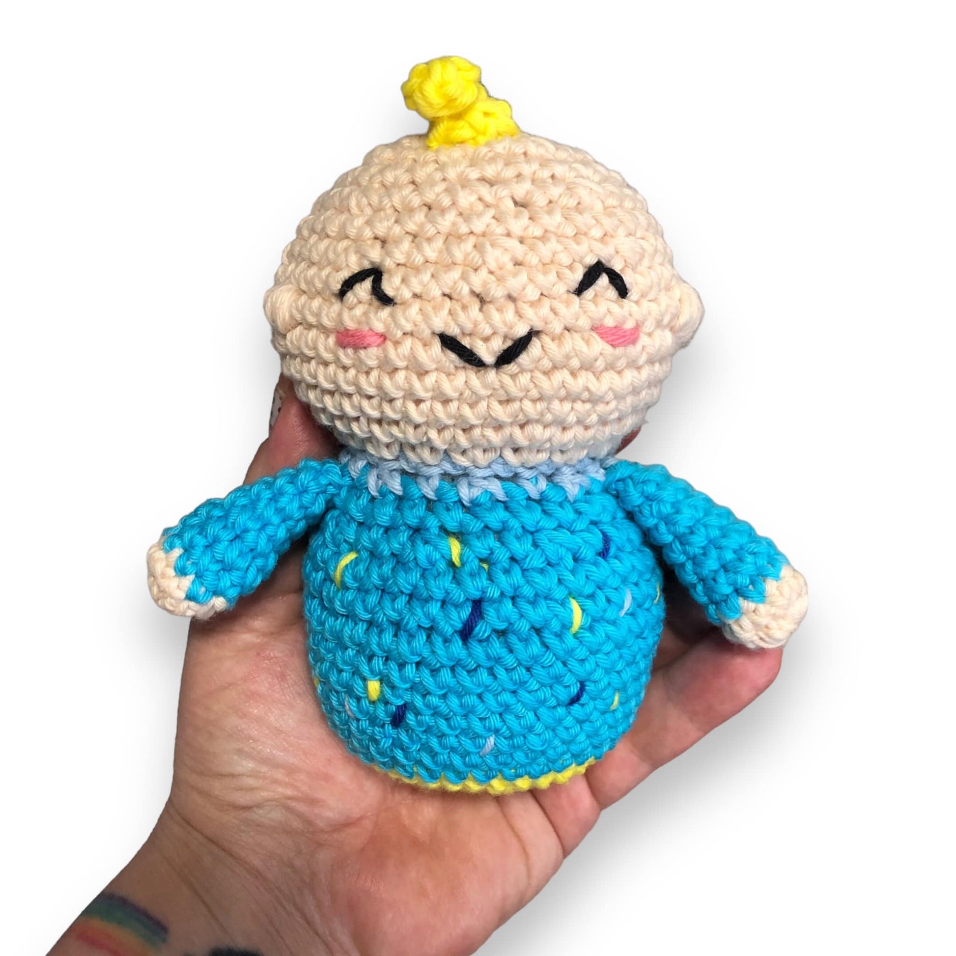 A crocheted doll resembling JJ from cocomelon being held up against a white background. 
