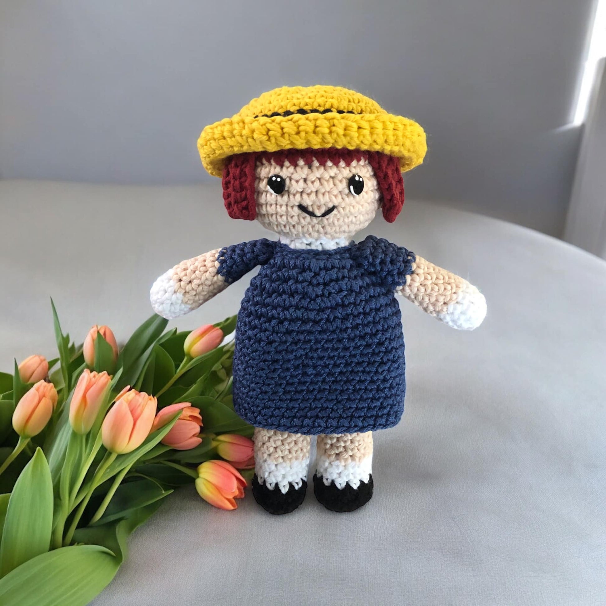 A crocheted doll of Madeline from the series Madeline standing next to a bouquet of pink tulips. 