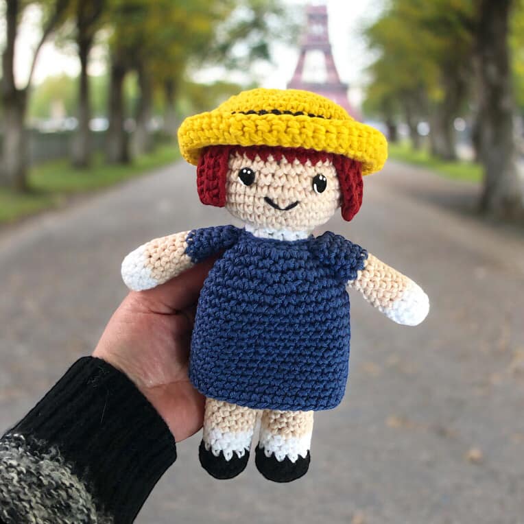 A crocheted doll resembling Madeline from the children's series Madeline being held up against a backdrop of a street in France featuring the Eiffel Tower.