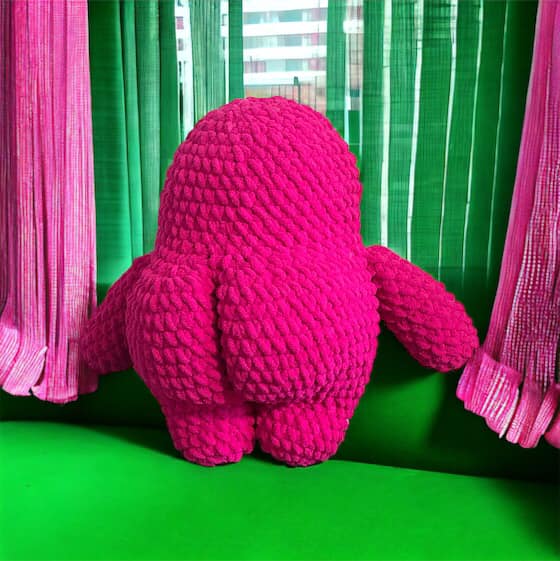 A crocheted pink monster plushie with a bubble butt, looking out a window and showing off the back side. 