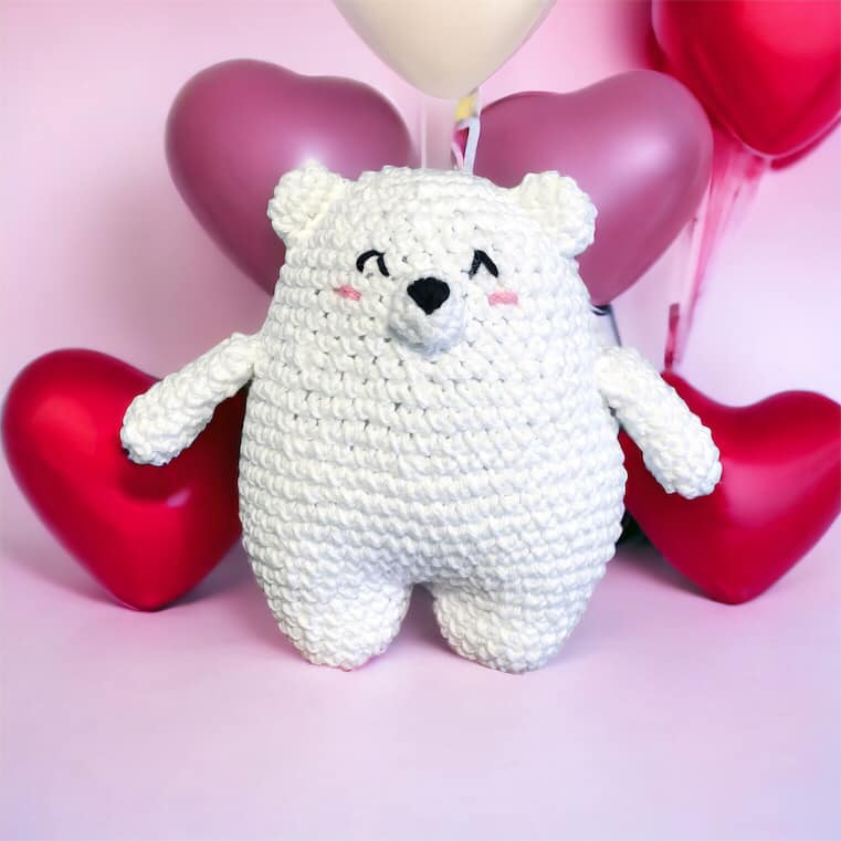 A crocheted doll resembling a polar bear standing against a pink background with heart balloons. 