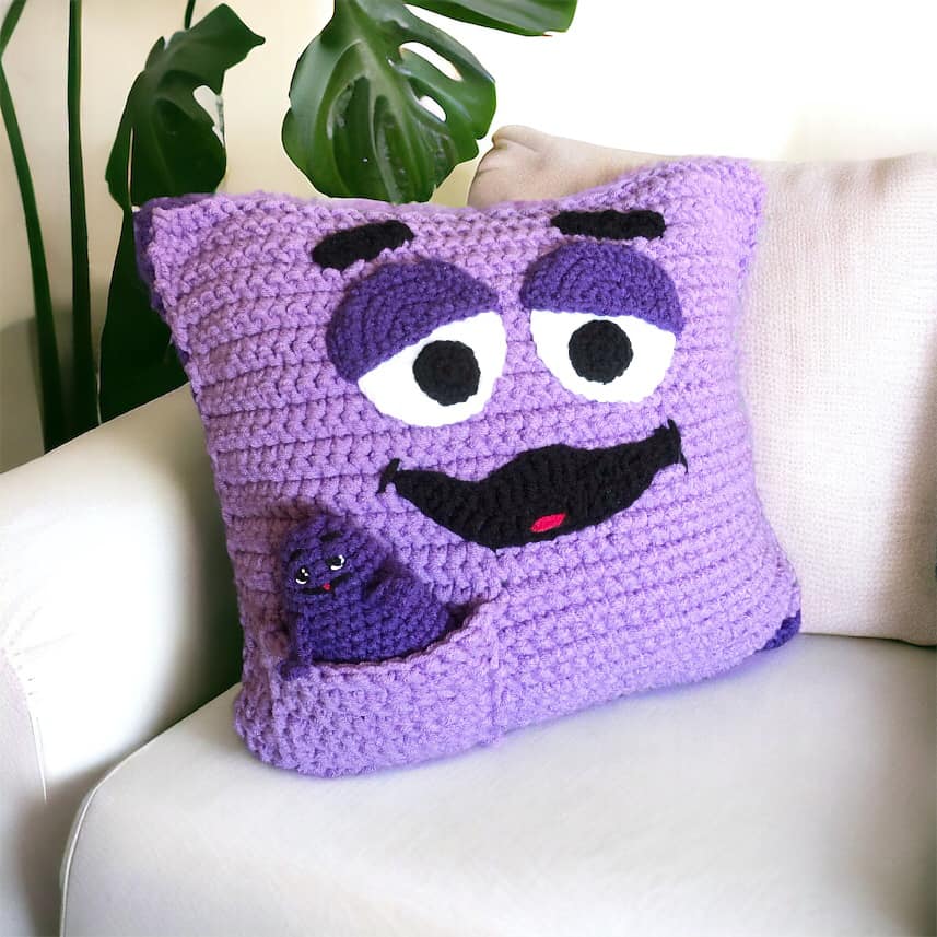 A crocheted Grimace pillow with a little crocheted Grimace doll in a little pocket sitting on a white coach. 