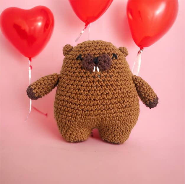 A crochet doll resembling a round beaver standing against a pink background with heart balloons. 