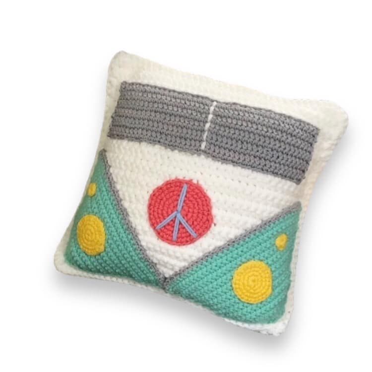 A crochet pillow resembling a 70s hippy van with a peace symbol on the front against a white background. 