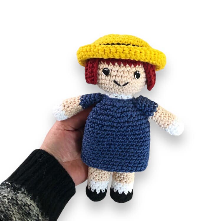 A crochet doll resembling Madeline from the series Madeline being held up against a white background. 