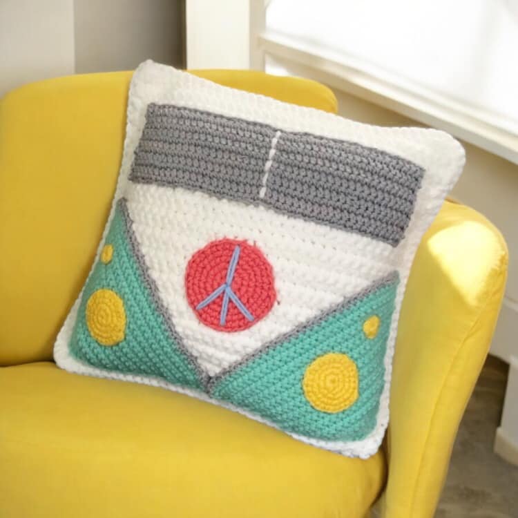 A crochet cushion with a van design, featuring peace symbols and multiple colors including white, grey, turquoise, coral, and yellow set against a yellow couch. 