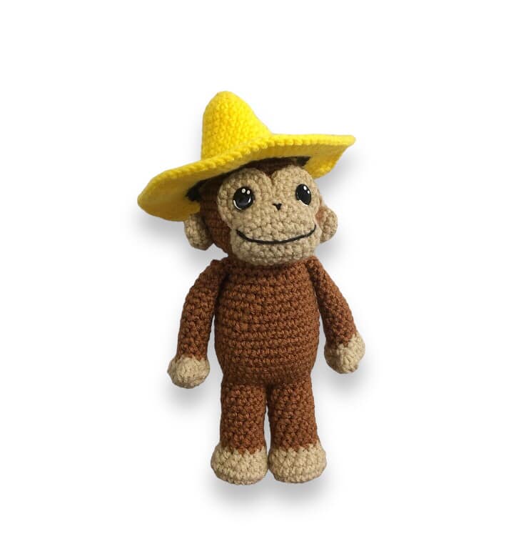A crocheted doll resembling Curious George wearing a crocheted yellow hat standing against a white background. 