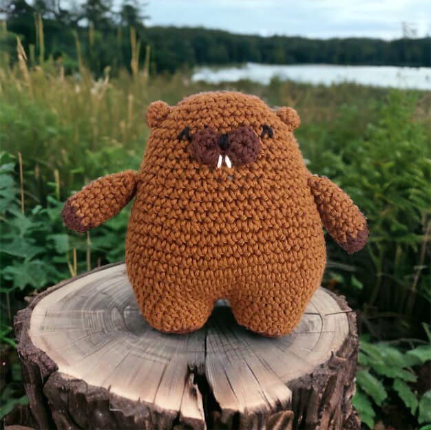 A crochet plushie of a beaver standing on a log near a lake and in a forested area. 