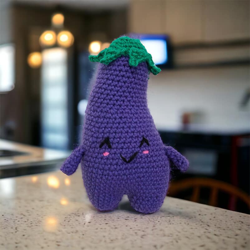 A crocheted doll resembling a cute eggplant standing on a white marble kitchen counter top. 