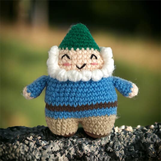 A crocheted doll of a smiling garden gnome with a green hat, blue jacket, beige pants, and a white beard standing on a rock. 