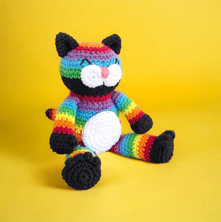 A crocheted rainbow cat doll sitting against a yellow background. 