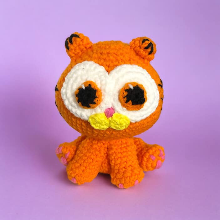 A crochet plush resembling garfield but as a kitten sitting down against a lavender background. 