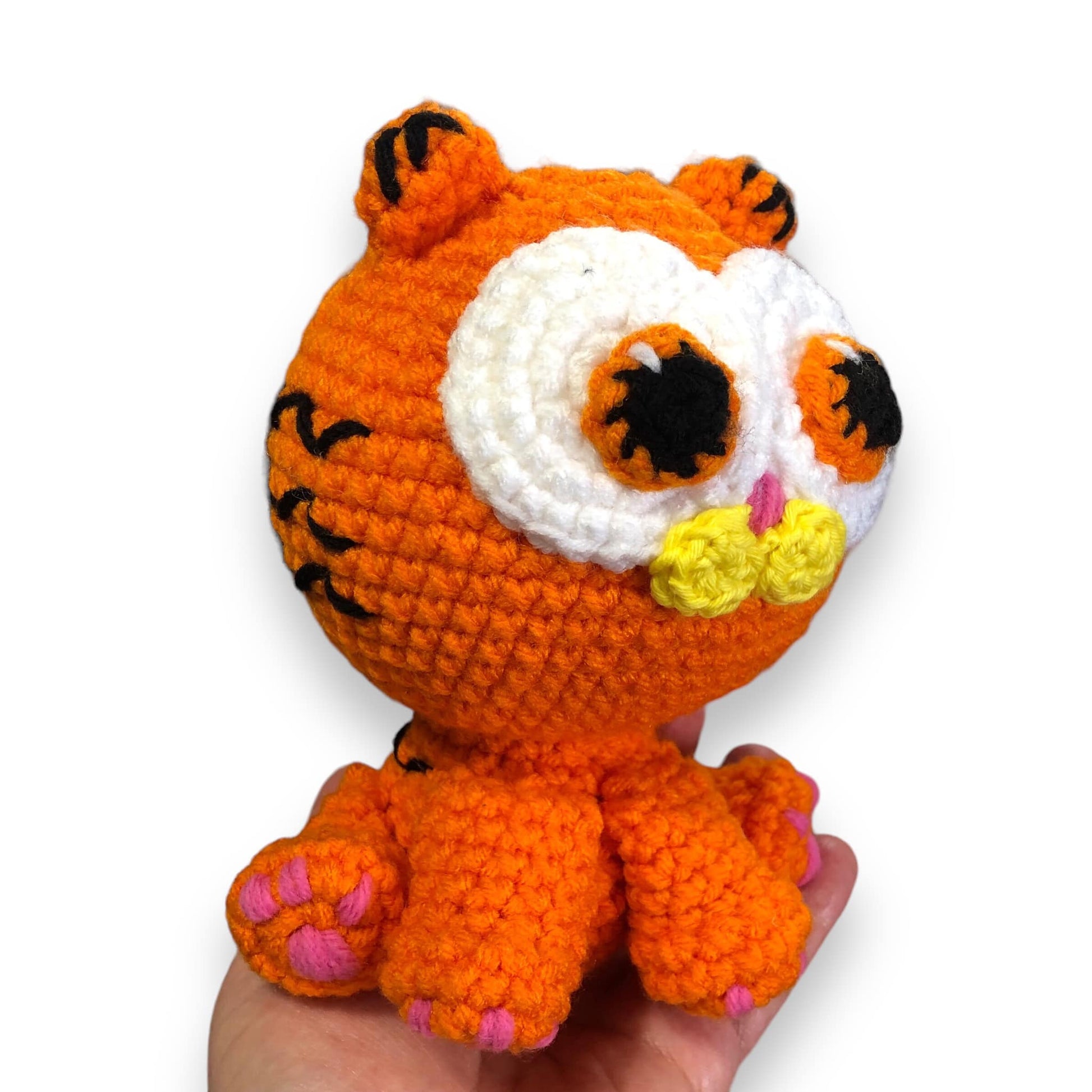 A crocheted doll of a kitten version of Garfield being held up by a hand showing off the right side of the face against a white background. 