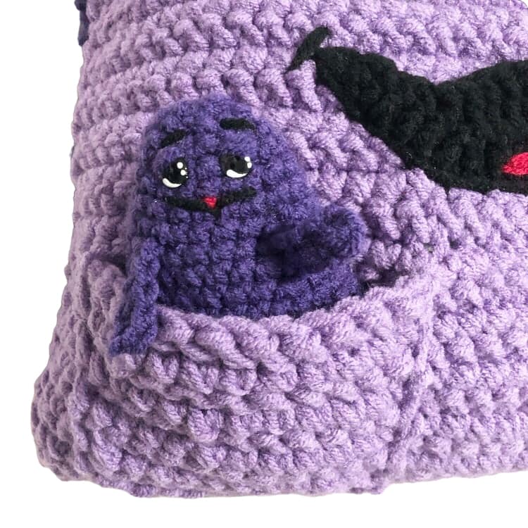 A crocheted doll of Grimace inside a pocket stitched onto a Grimace pillow. 