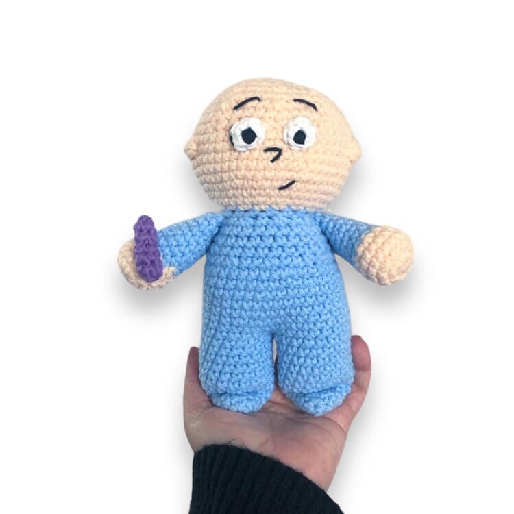A crocheted doll resembling Harold from Harold and the Purple Crayon being held up against a white background. 