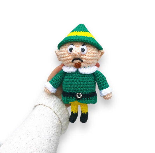 A crocheted version of the Head Elf from Rudolph the Red Nosed Reindeer being held up against a white background. 