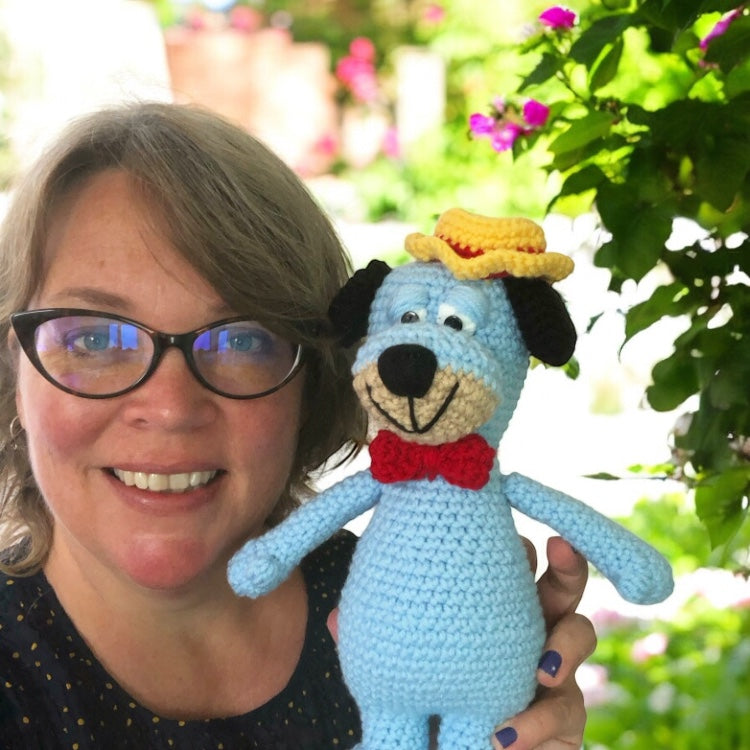 A smiling woman holding up a crochet version of Huckleberry Hound against the background of a garden. 