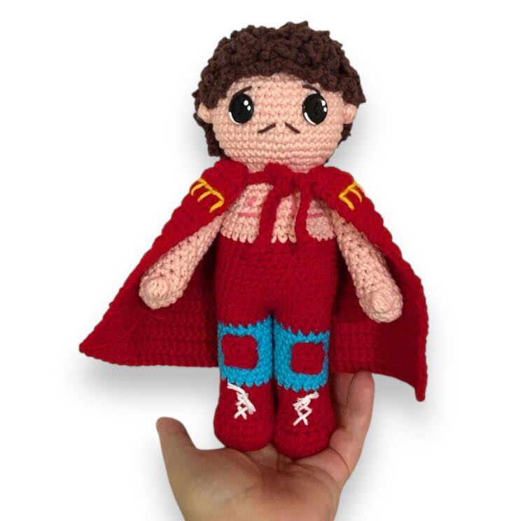 Crocheted doll of Ignacio from Nacho Libre with cape and boots being held against a white background. 