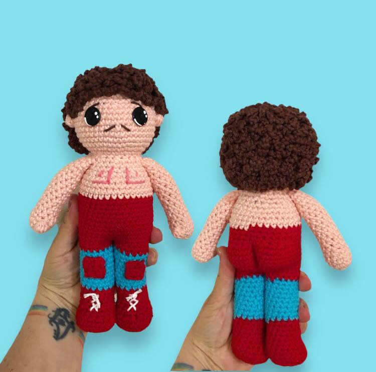 Crocheted doll of Ignacio from Nacho Libre with cape and boots held against a blue background. 