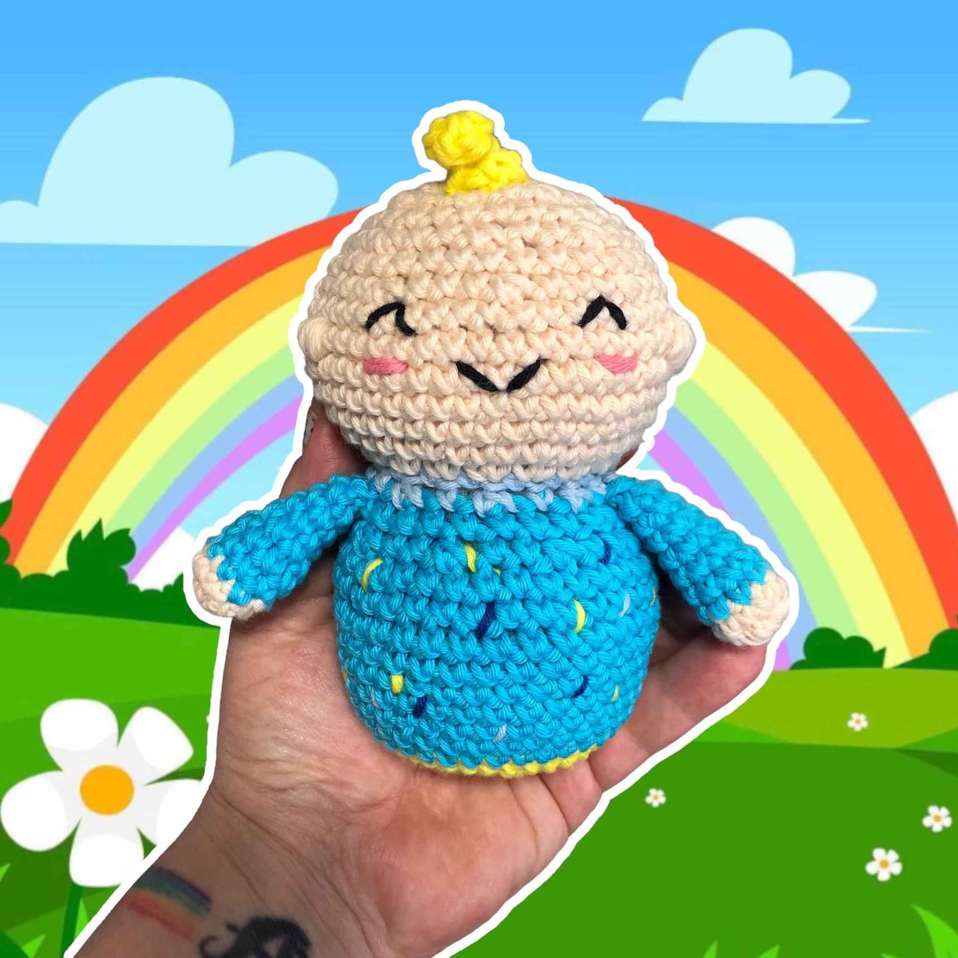 A crochet doll resmbling JJ from Cococmelon being held up against a background with a rainbow and grassy fields with daisies. 