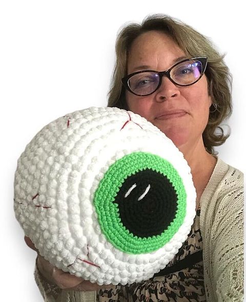 A woman holding up a giant crochet eyeball with a green iris. 