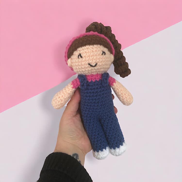 A crocheted doll resembling the kids content creator Ms. Rachel being held up against a pink and white background. 