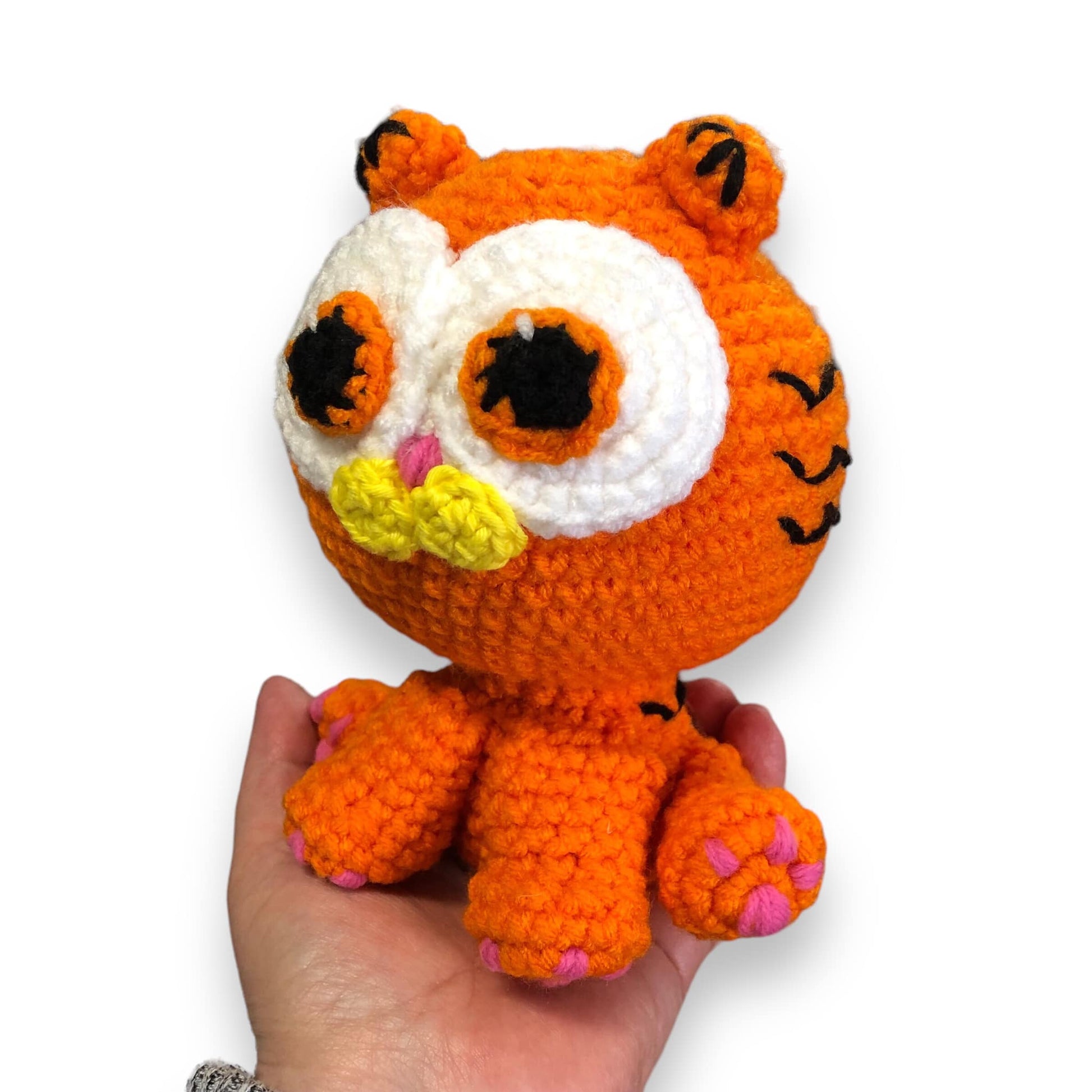 A crocheted doll of a kitten version of Garfield being held up by a hand showing off the left side of the face against a white background. 